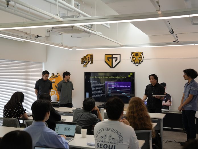 Students sitting in a classroom and watching other students do a presentation using a big-screen monitor.