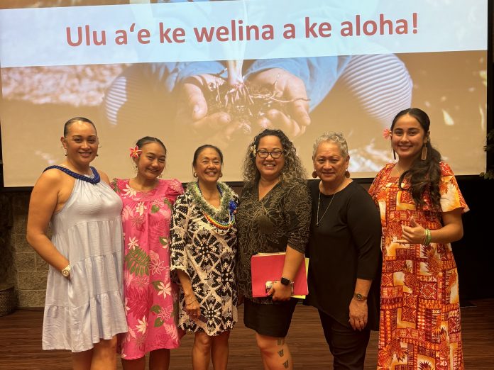 A group of women smiling adn standing in front of a large screen that says: Ulu a‘e ke welina a ke aloha!