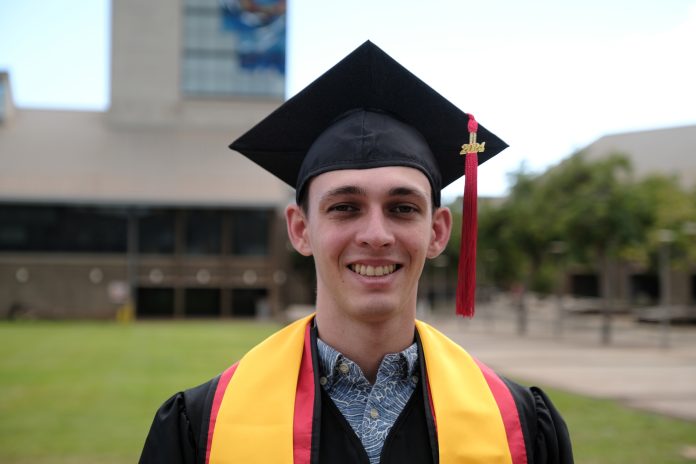 A man wearing a graduation cap and gown and standing outdoors with the UH West Oahu library tower in the background.