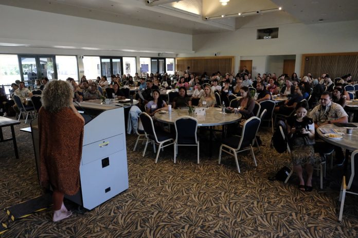 The back of a woman speaking at a podium to a large audience seated at round tables in a conference room.