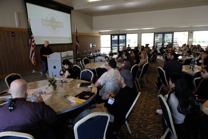 A man standing at a podium in a room full of men and women sitting at round tables.