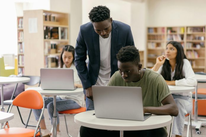 A teacher looking over a student sitting at a desk and typing on his laptop in a classroom. There are two other students in the background.