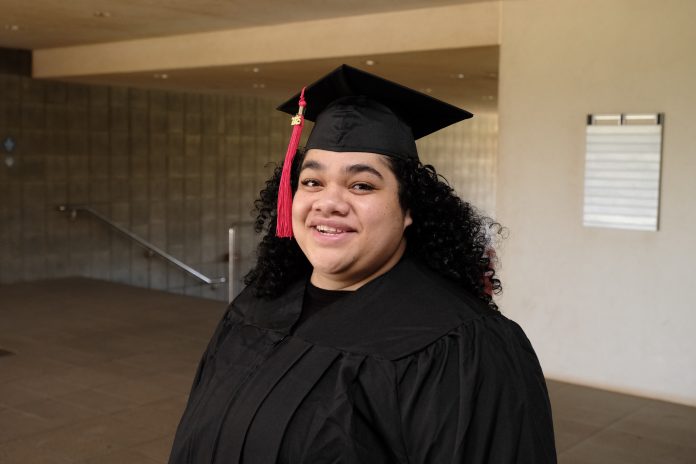 A woman wearing a cap and gown, and standing in a breezeway.