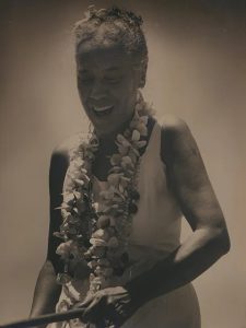 A sepia toned image of ʻIolani Luahine with a lei around her neck, her hair in a bun, and a smile on her face.