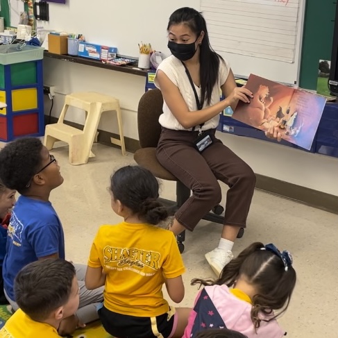 A teacher sitting in front of a classroom and reading a book to students seated on the floor in front of her.