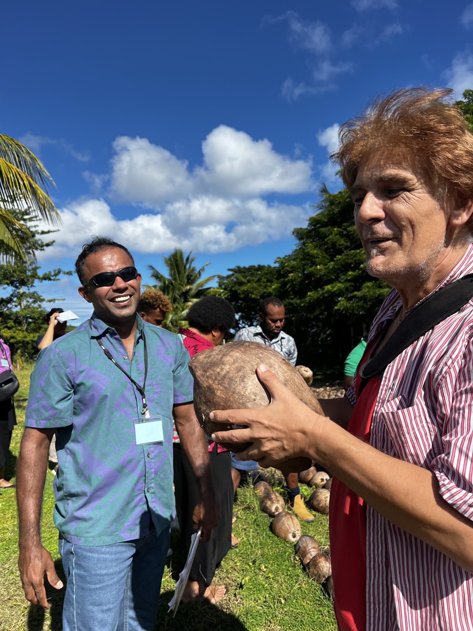 Two men chatting outdoors and one is holding a coconut.