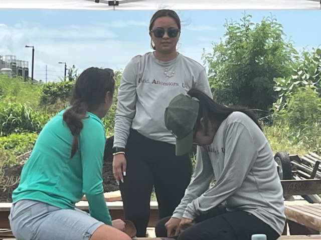 An outdoor photo of two people sitting while facing each other, and another person standing by them.