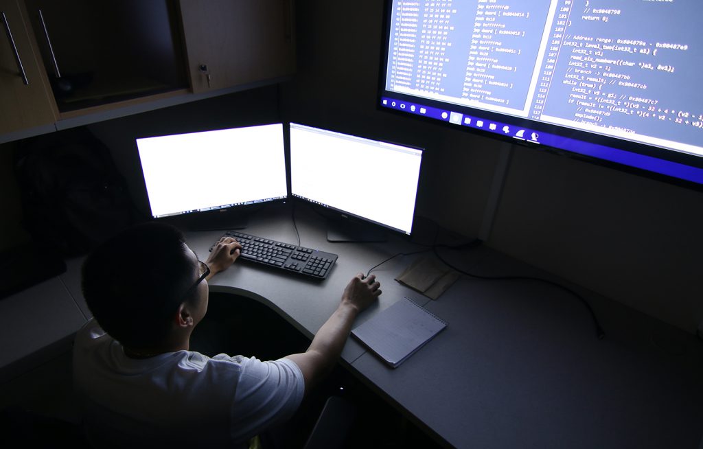 Computer student in front of 3 monitors in a dark room.