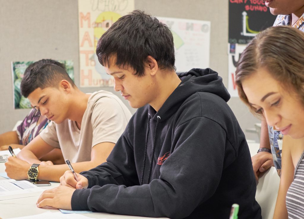 3 students sitting on a long table looking down at their assignments.