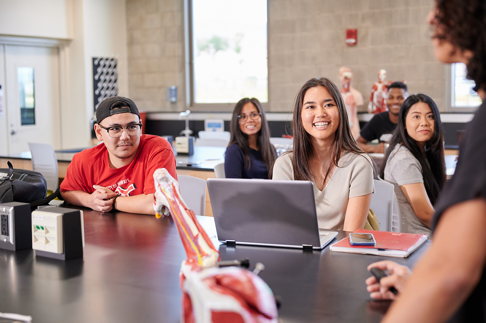 Five students sitting and smiling while listening to a professor presenting.