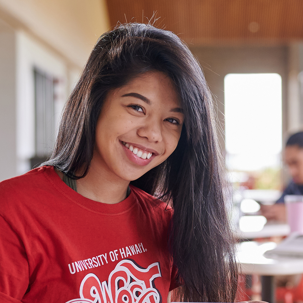 Close-up Of A Student Smiling While Sitting At A Table In The Hallway Wearing A Red T-shirt.