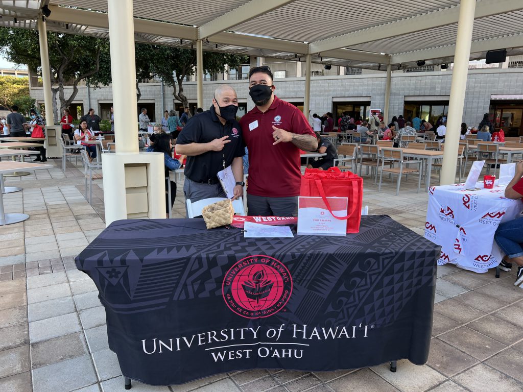 Two men pose in front of a table with a "UH West Oahu" logo
