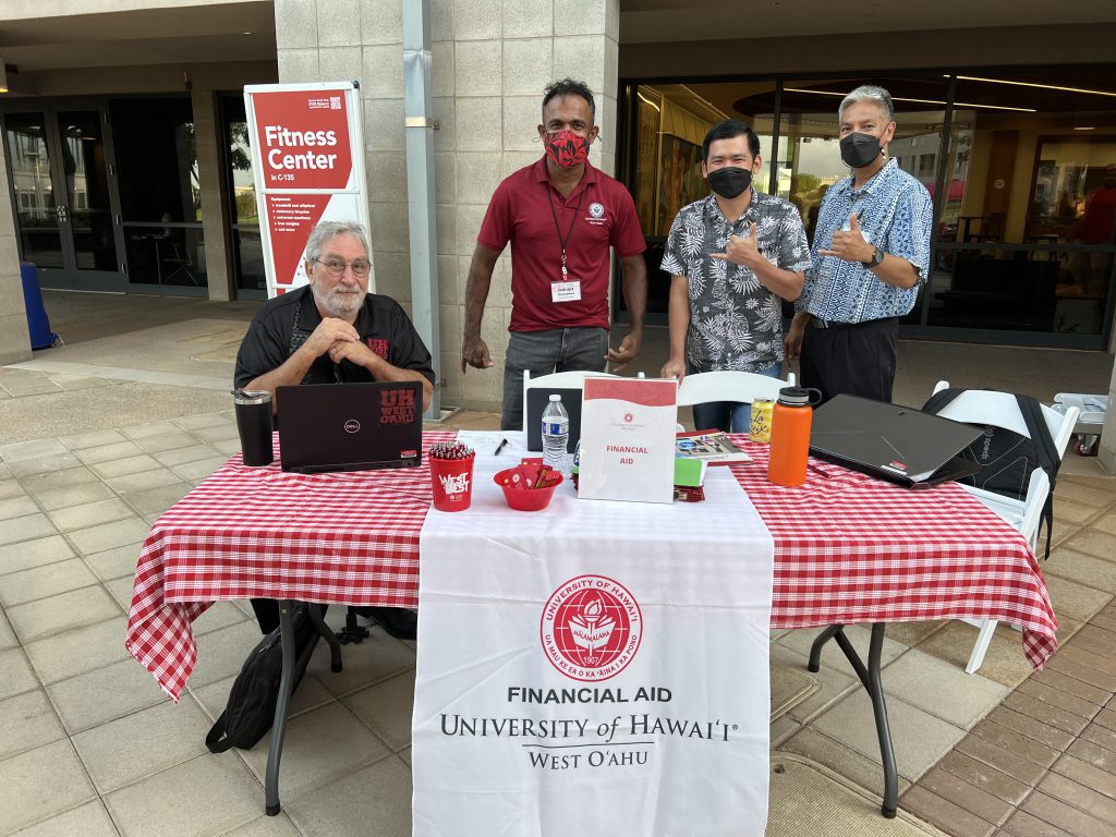 Four men sit and stand behind a table promoting University of Hawaii-West Oahu.