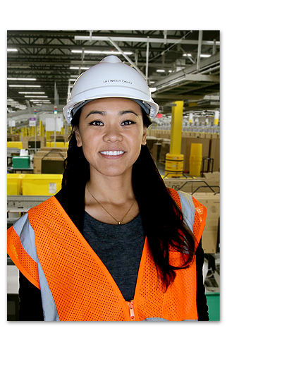 Student wearing a white hard hat and orange safety vest in a warehouse. This image's background has been modified.